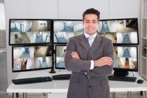 A man with crossed arms stands in front of monitors showing camera footage.